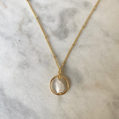Gold necklace with a circular pendant featuring a white stone on a marble surface