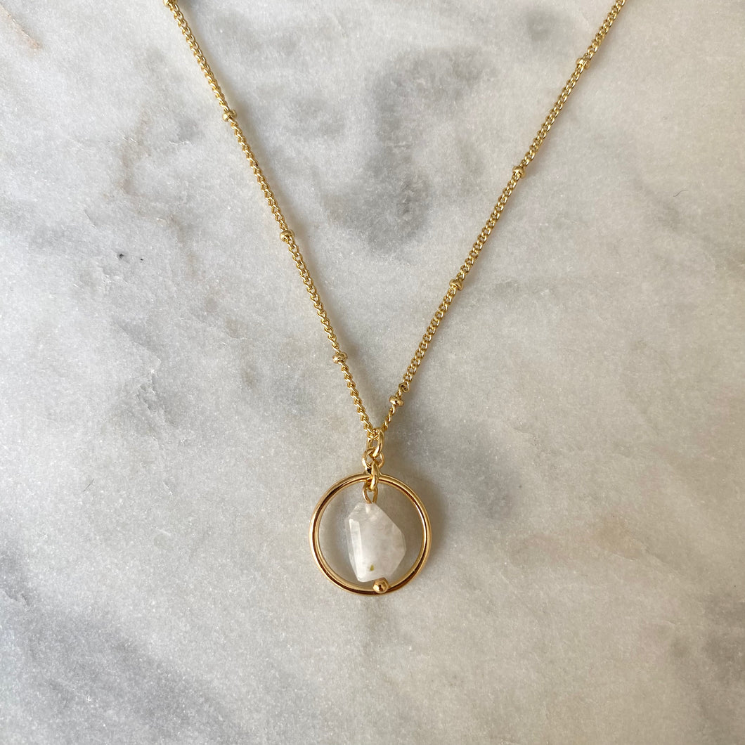 Gold necklace with a circular pendant featuring a white stone on a marble surface