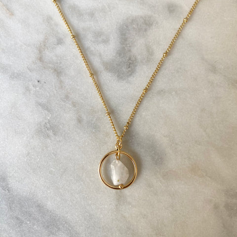 Gold necklace with a circular pendant featuring a white stone on a marble surface